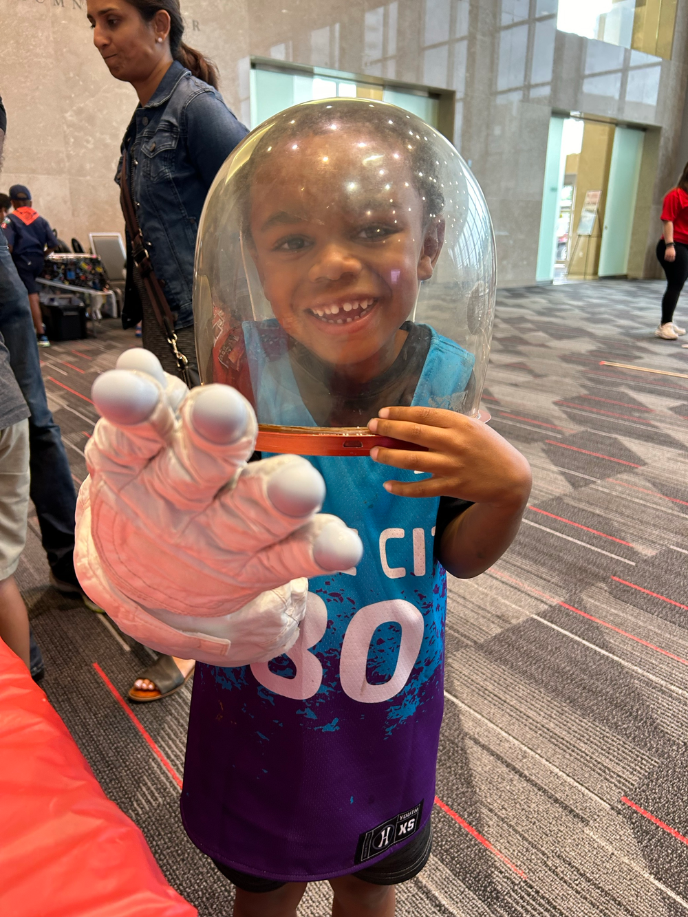 A young boy in a blue jersey smiles while wearing an astronaut space glove and helmet. 